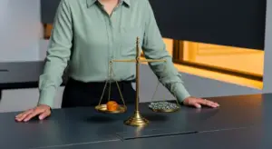 Gold balance scales on a desk with an orange gemstone on the left pan and a pile of coins on the right, a person in a green shirt standing behind them.
