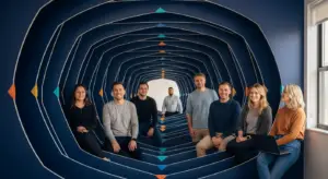 Nine coworkers sit along curved blue benches in a vivid blue geometric tunnel mural, posing for a team photo in a bright office.
